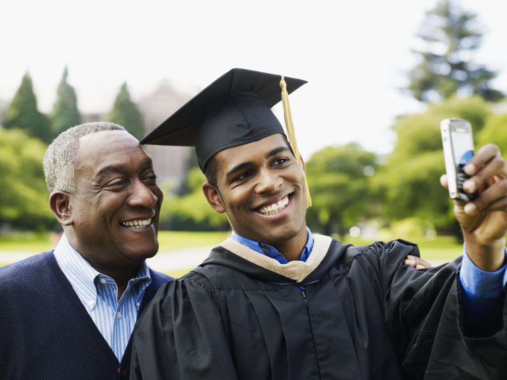 Young man in graduation gown taking self-portrait photograph with father on campus