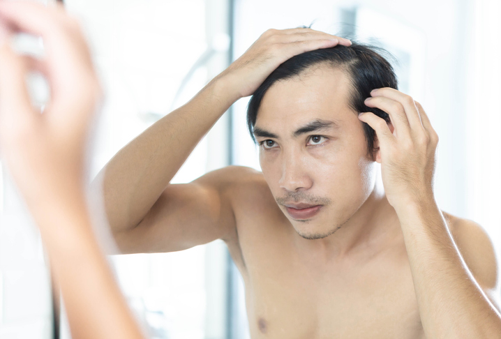 Mid Adult Man Looking In Mirror While Touching His Receding Hairline At Bathroom