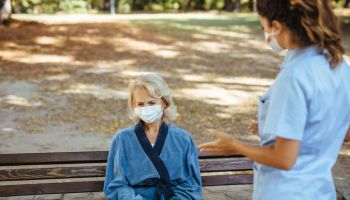 Elderly woman with protective face masks on sitting on a bench and talking to healthcare worker at nursing home