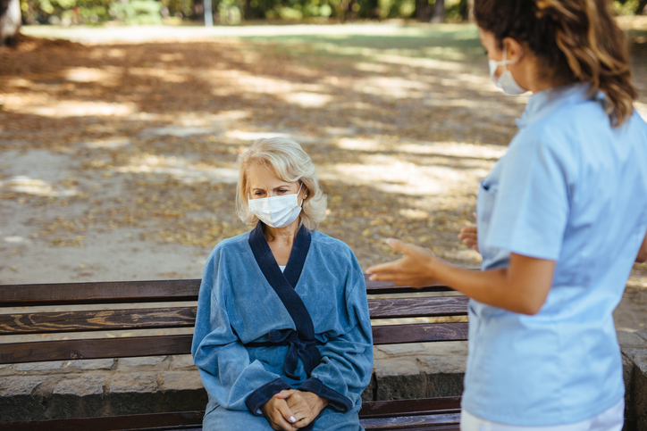 Elderly woman with protective face masks on sitting on a bench and talking to healthcare worker at nursing home