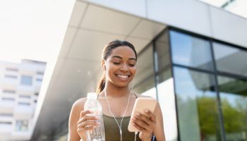 Black woman looking at cellphone, listening to music in earphones, choosing playlist, holding water on training outdoors