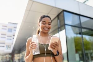 Black woman looking at cellphone, listening to music in earphones, choosing playlist, holding water on training outdoors