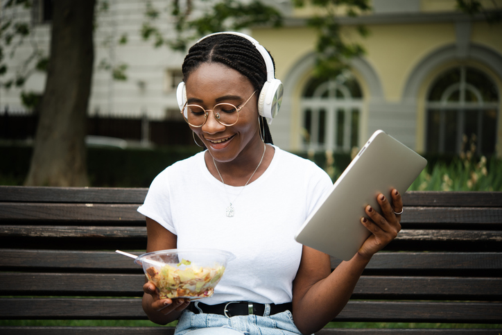Portrait of a young African woman eating, using tablet and enjoying music outdoors