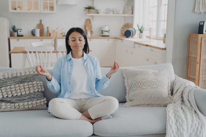 Serene woman practices yoga, meditates on couch in lotus position. Breathing exercises for mental balance, anxiety relief. Healthy lifestyle at home.