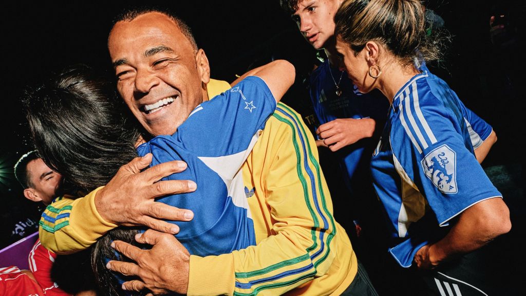 A smiling older man in a yellow and blue sports jersey embracing several people, including a woman in a blue jersey, in what appears to be a celebratory or joyful moment.
