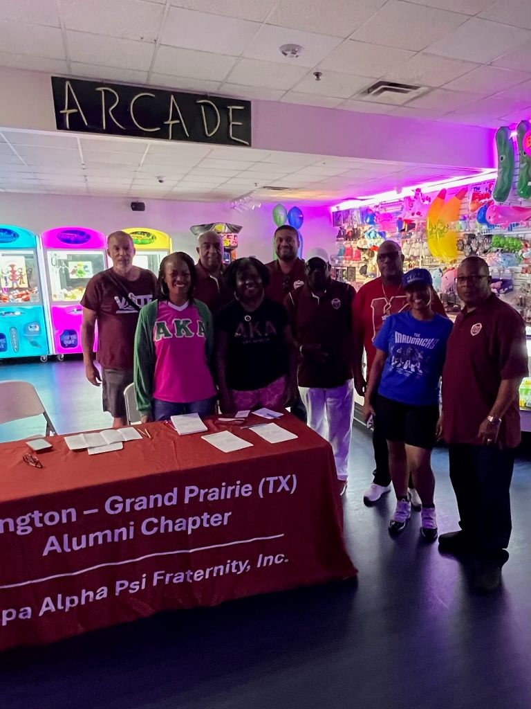 A group of people wearing Alpha Psi Fraternity apparel standing in front of an "Arcade" sign, with a table displaying the Washington-Grand Prairie (TX) Alumni Chapter of the fraternity.