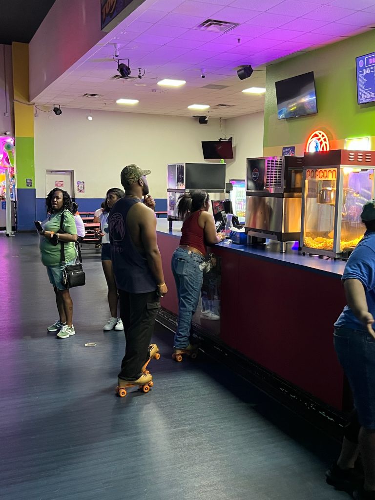 A group of people in a colorful, neon-lit entertainment venue, some wearing roller skates, standing in front of a concession counter with a popcorn machine.