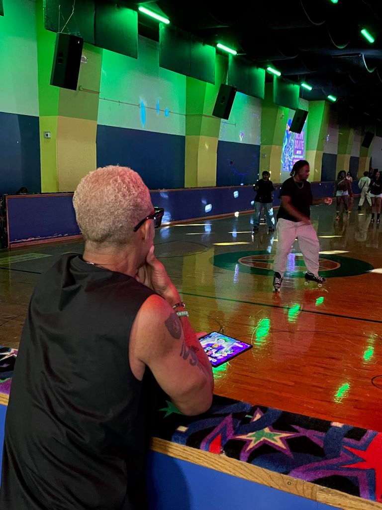 A person in a black shirt and shorts stands on a brightly lit indoor basketball court, holding a mobile device and surrounded by colorful walls and lighting.