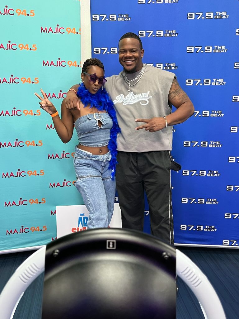 Two people posing in front of MAJIC 94.5 radio station branding, one wearing a blue feather boa and the other wearing a gray t-shirt with "Donnell" printed on it.