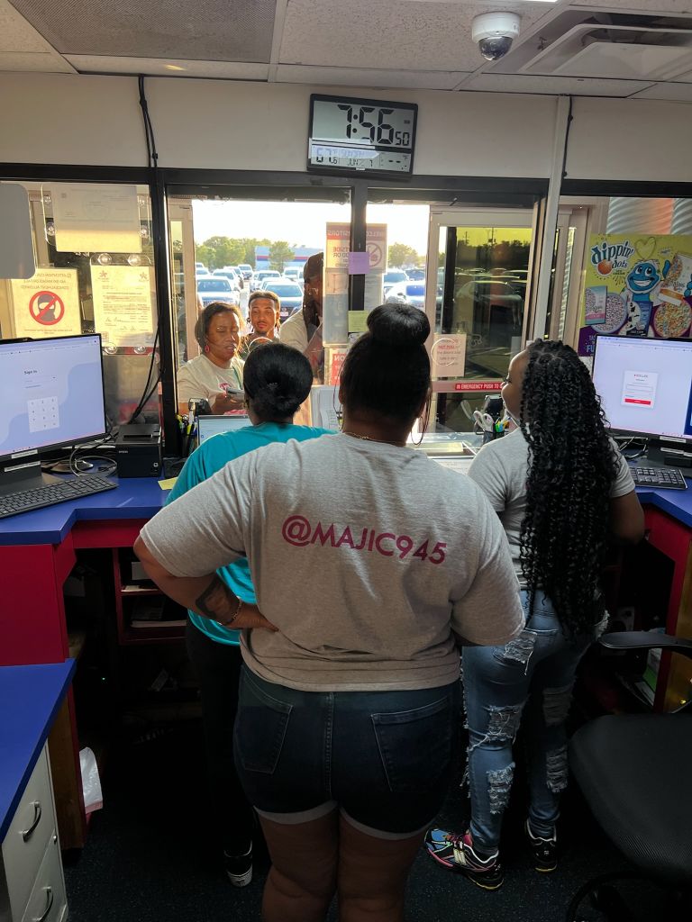A group of people standing in front of a convenience store counter, with a digital clock display showing 7:56. One person is wearing a shirt with the text "@MAJIC945".