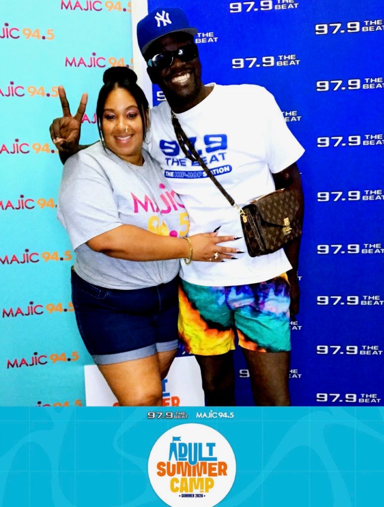 Two people posing for a photo in front of Majic 94.5 radio station branding, one wearing a Majic 94.5 t-shirt and the other a NY Yankees hat.