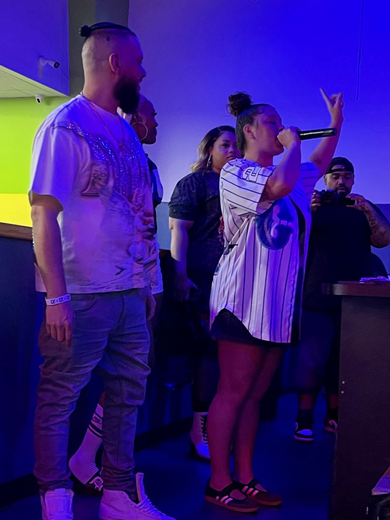 A group of people performing on stage, with a person in a white jersey singing into a microphone and others standing behind them.