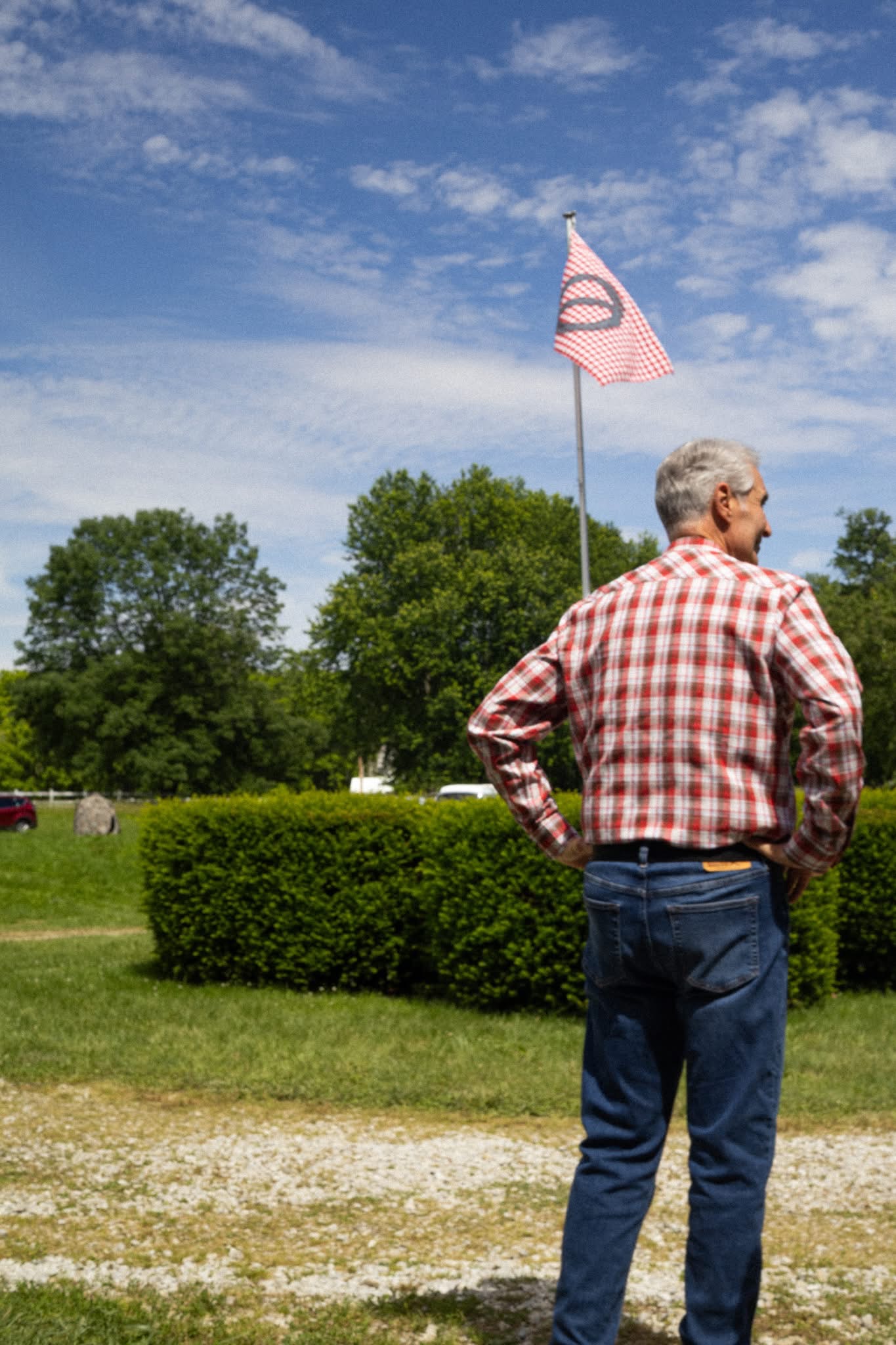 An older man in a plaid shirt stands facing a red-and-white checkered flag on a pole, with a grassy field and trees in the background.