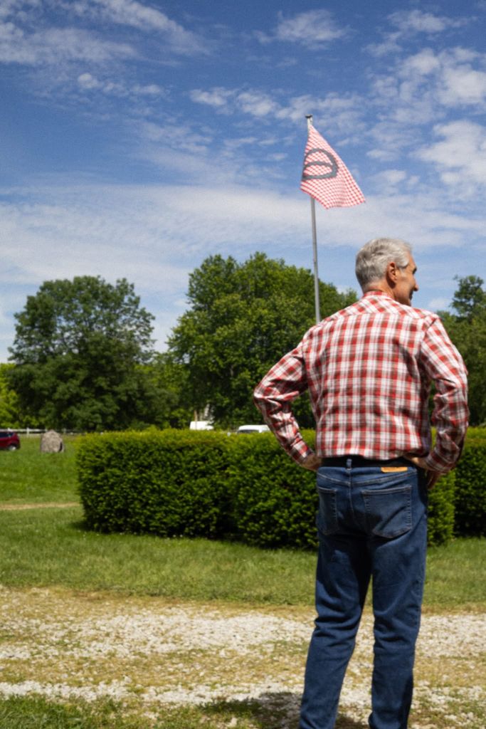 An older man in a plaid shirt stands facing a red-and-white checkered flag on a pole, with a grassy field and trees in the background.