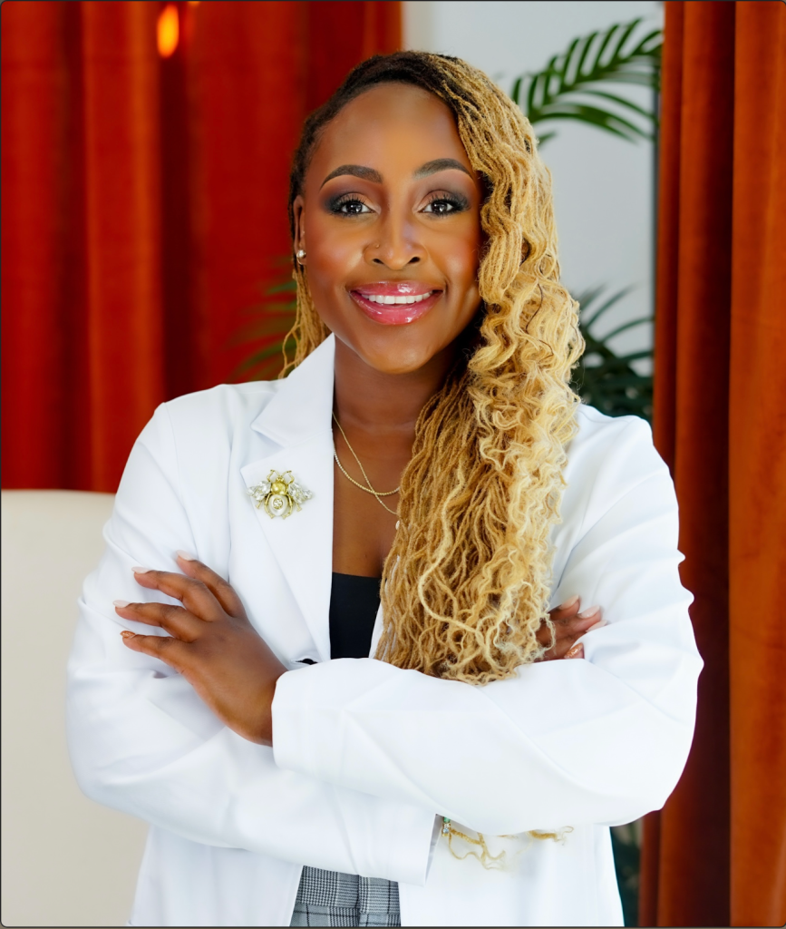 A smiling woman with curly blonde hair wearing a white blazer and standing in front of a tropical plant backdrop.