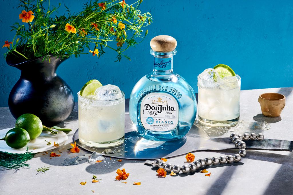 A still life featuring a bottle of Don Julio Blanco tequila, a glass of margarita, limes, and a vase of colorful flowers on a blue background.