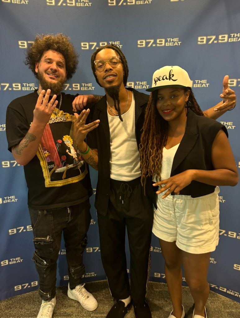 Three people posing in front of a backdrop with "97.9 THE BEAT" branding, one wearing a hat that says "Speak".