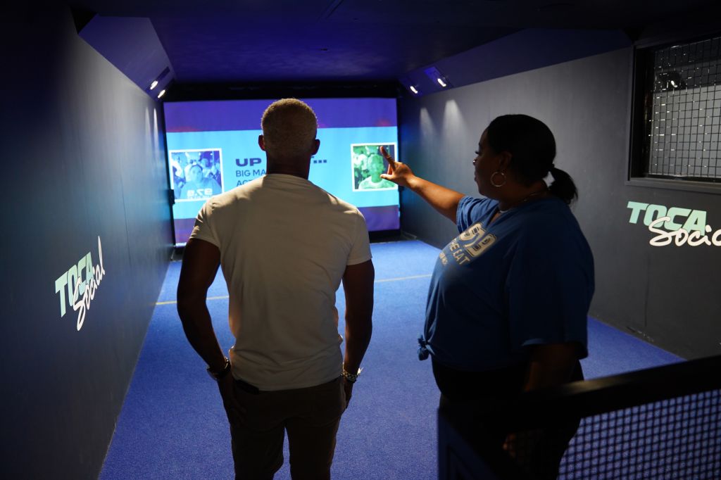 Two people, one pointing at a large screen displaying "TOCA SOCIAL" and "UP BIG MATCH" branding.