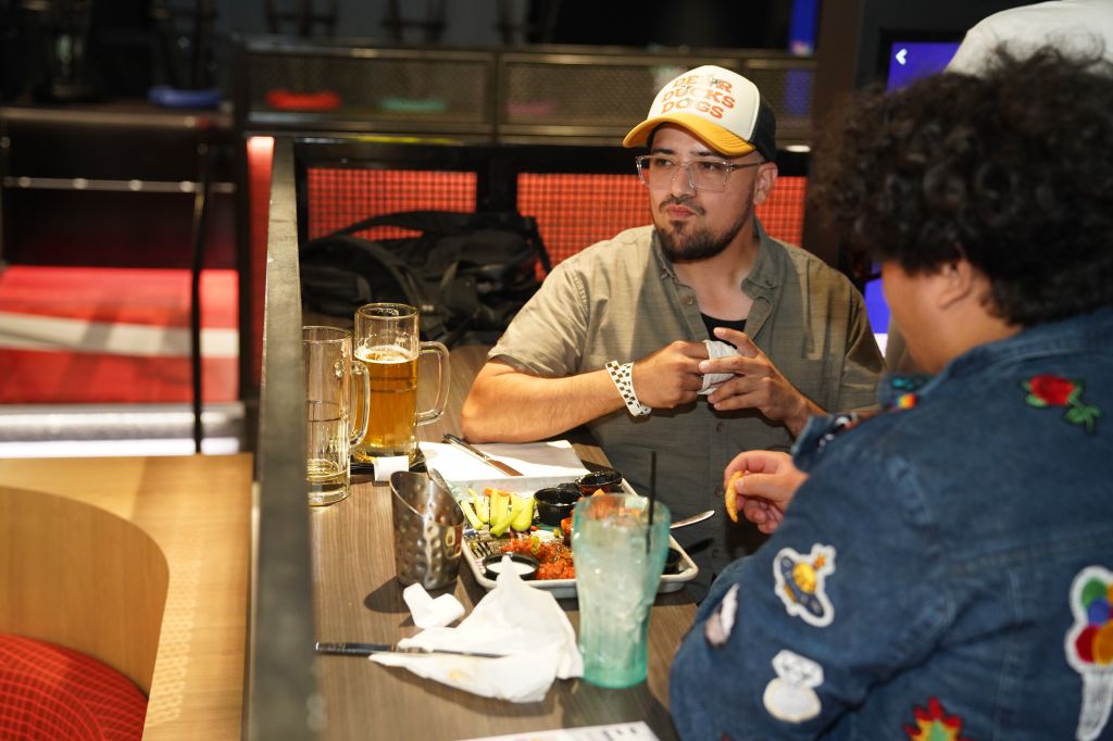 Two men sitting at a bar, one wearing a yellow baseball cap and drinking a beer, the other wearing a denim jacket with patches.