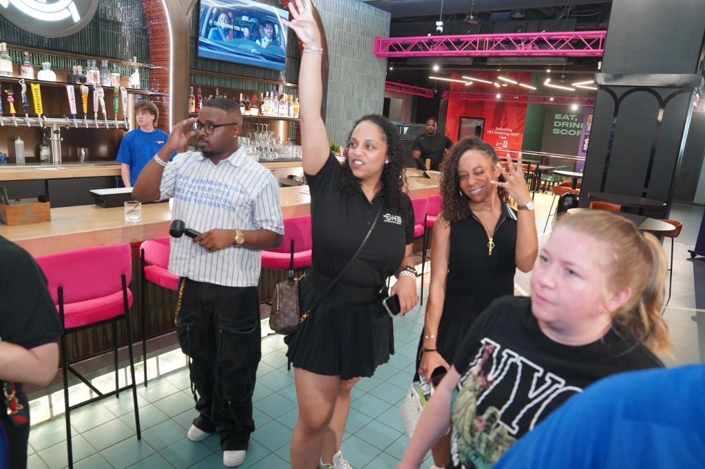 A group of people, including a man and several women, standing in a bar or restaurant setting with pink and black decor and a TV screen visible.