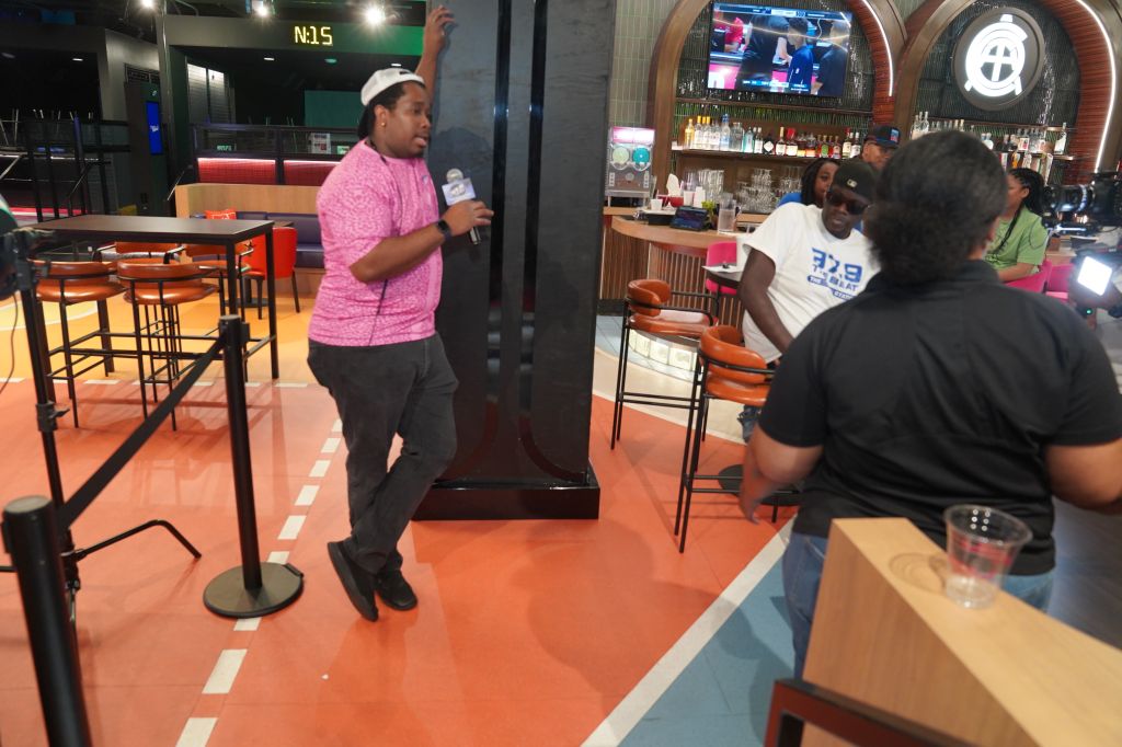 A person wearing a pink shirt standing in a sports bar or restaurant, with other people visible in the background.