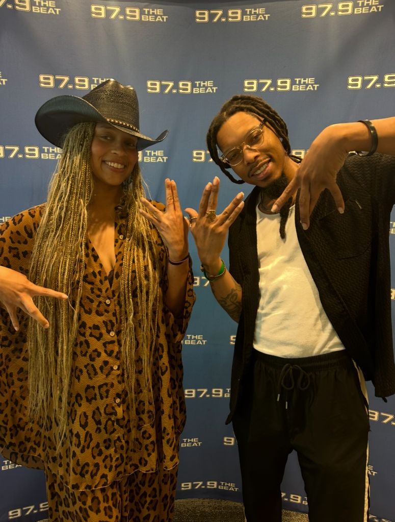 Two people posing in front of a backdrop with "97.9 The Beat" branding, one wearing a leopard print outfit and cowboy hat, the other wearing sunglasses and a casual outfit.