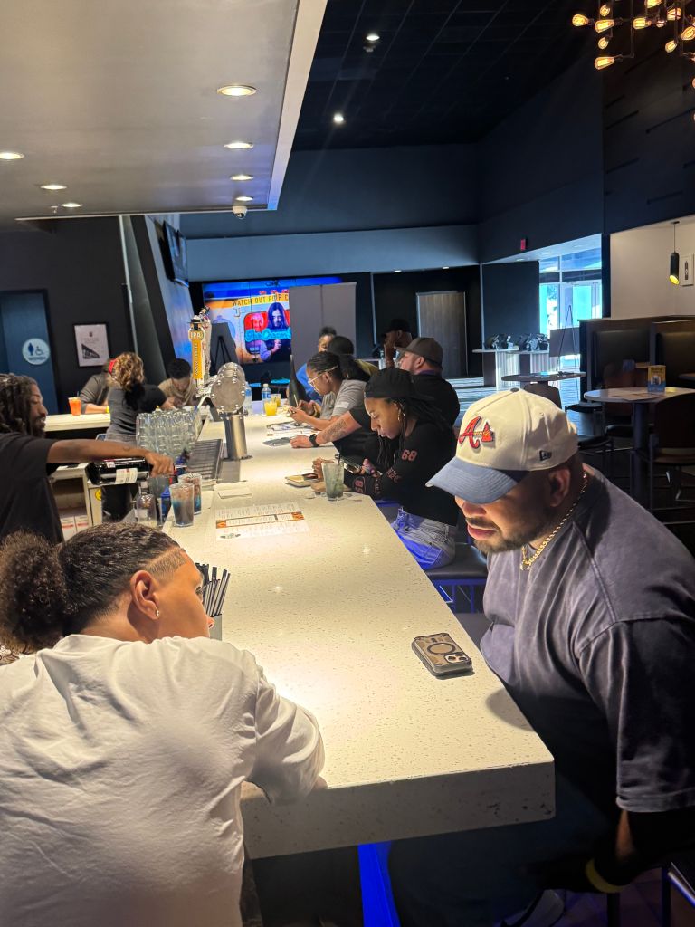 A group of people sitting at a bar counter, watching a TV screen displaying a message. The bar has a dark, modern decor with pendant lights overhead.