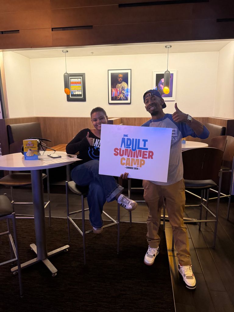 Two people sitting at a table, one holding a sign that says "Adult Summer Camp Summer 2023".
