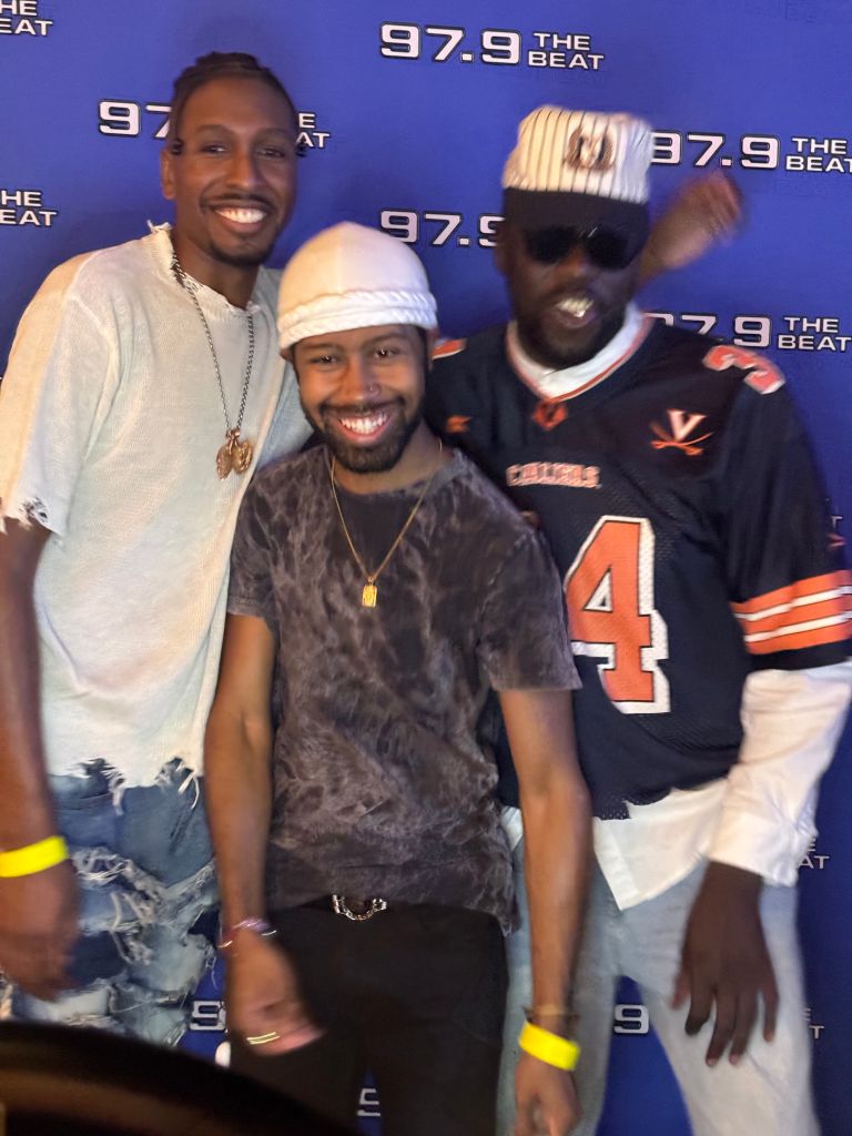 Three smiling men posing in front of "97.9 The Beat" backdrop, wearing casual clothing and accessories.