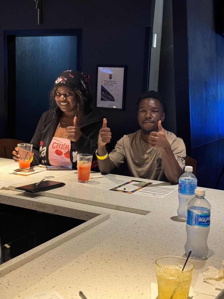 Two people sitting at a table, smiling and giving thumbs up. They have drinks and snacks in front of them.