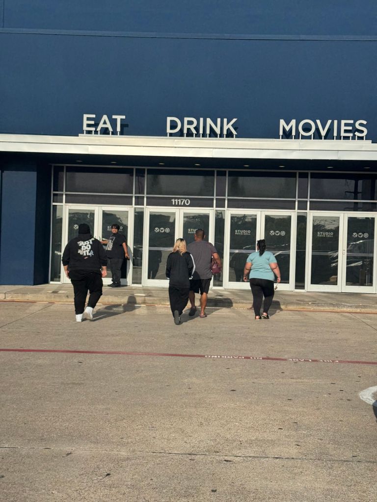 Exterior of a building with the text "EAT DRINK MOVIES" displayed prominently. Several people are walking outside the entrance.
