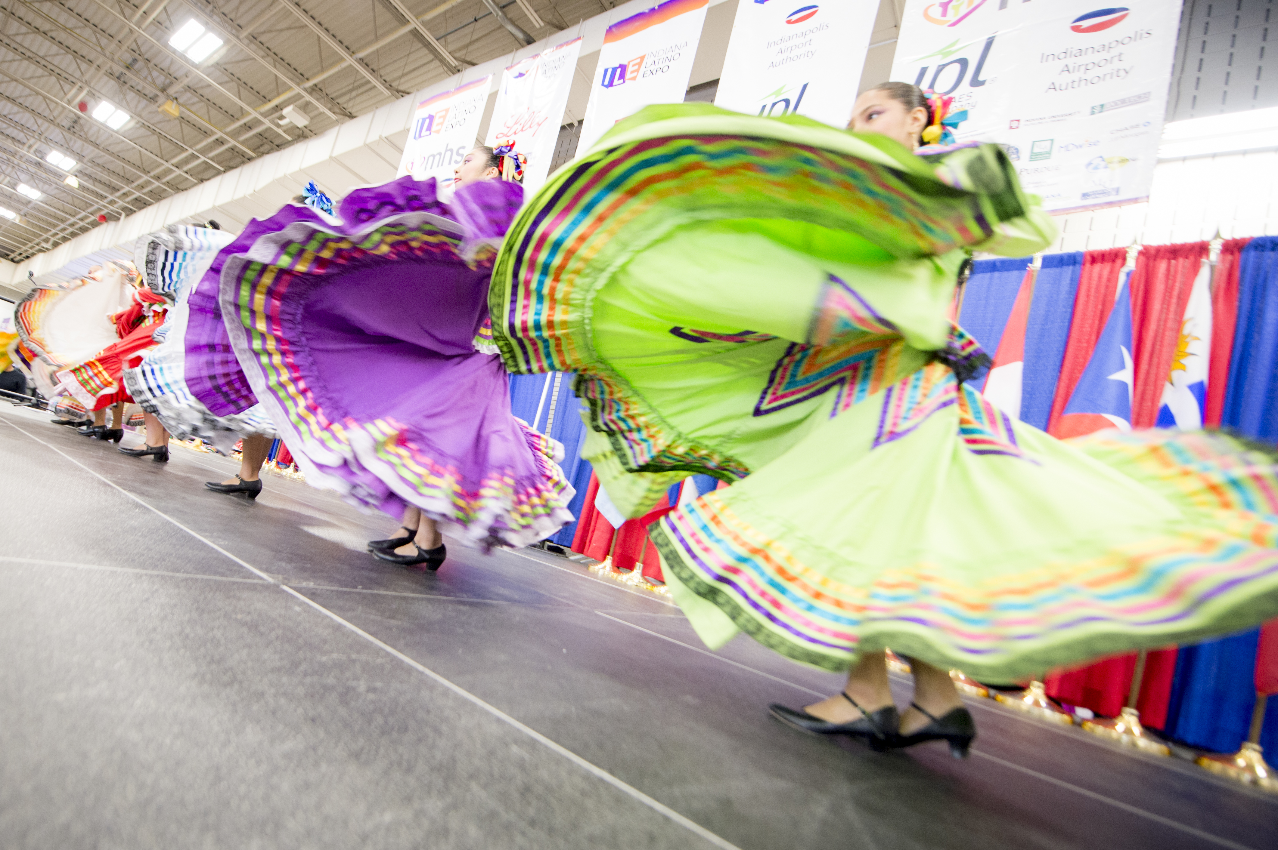 Colorful, flowing dresses of performers twirling on stage at an event or festival, with banners and signage visible in the background.
