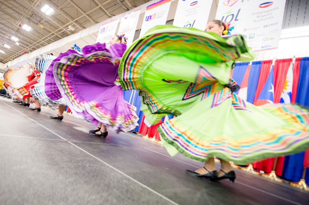 Colorful, flowing dresses of performers twirling on stage at an event or festival, with banners and signage visible in the background.
