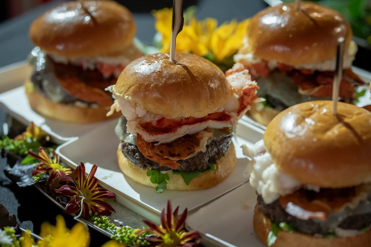 Closeup of several gourmet hamburger sliders with lettuce, tomato, and other toppings, arranged on a plate with colorful flowers.